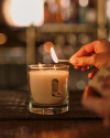 Hand lighting a candle in a glass holder with a cowboy boot design, placed on a wooden surface.