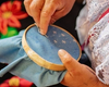 A person embroiders blue fabric with white thread using a needle and hoop. Red flowers are visible in the background.