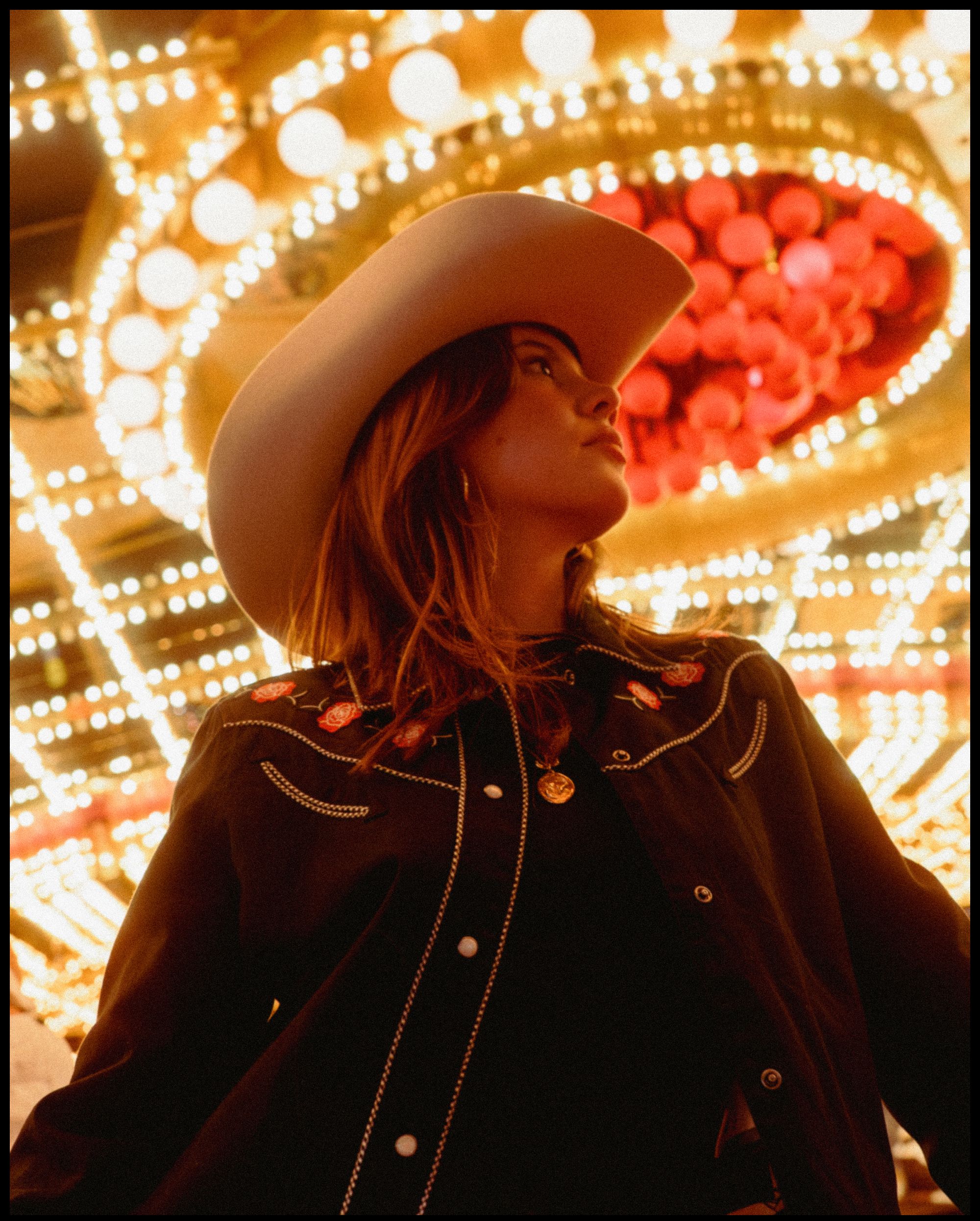A person in a cowboy hat and embroidered jacket stands under bright, round lights and red decorations, looking to the side.