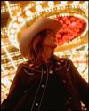 A person in a cowboy hat and embroidered jacket stands under bright, round lights and red decorations, looking to the side.