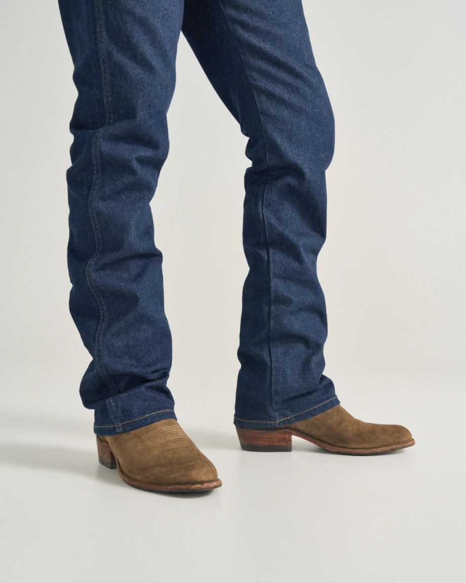 The Johnny in Gravel Suede. Wearing blue jeans and brown Gravel Suede cowboy boots, the person stands on a light-colored surface. Only their legs and feet are visible.