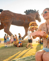 Woman enjoying a large burger at an outdoor festival, with a giant horse sculpture and crowd in the background.