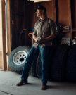 A man in a cap, button-up shirt, jeans, and boots stands in a garage next to four stacked tires, looking toward a source of light.