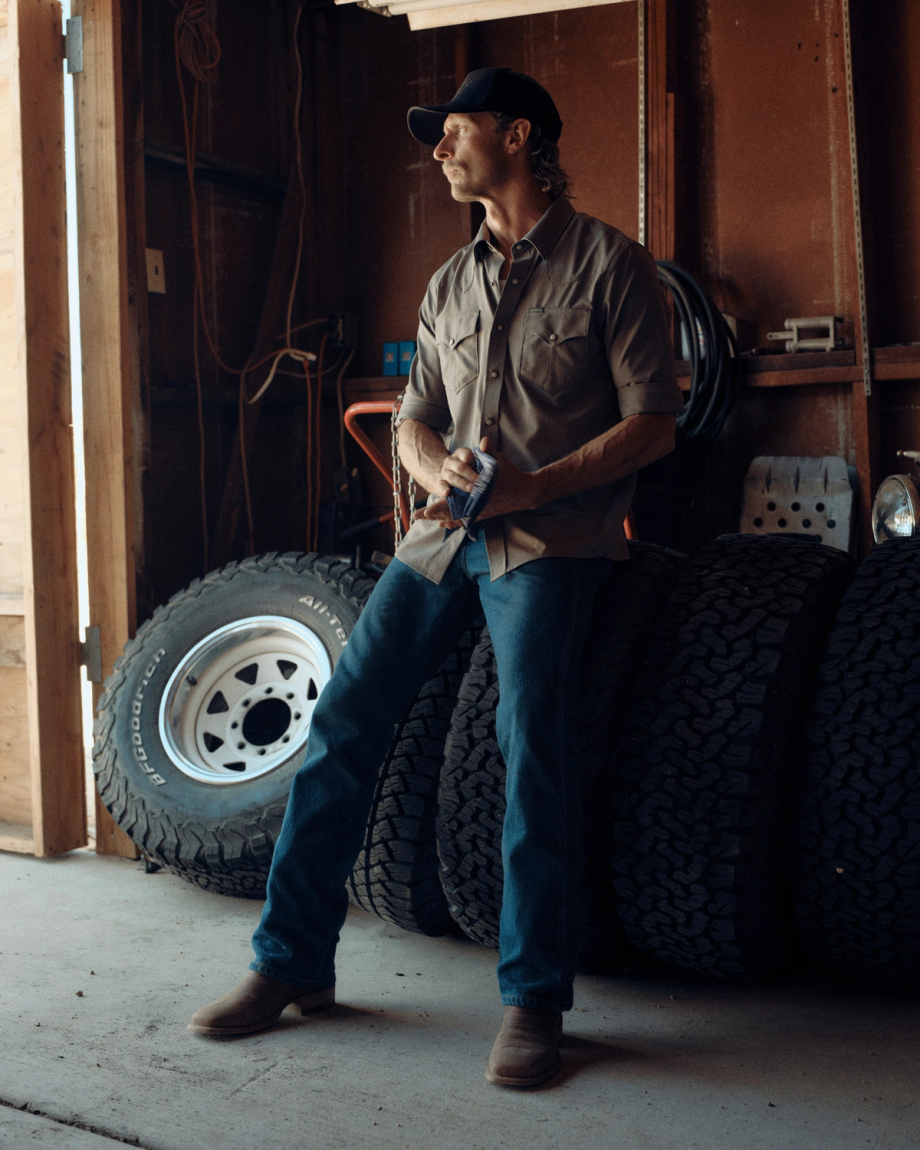 A man in a cap, button-up shirt, jeans, and boots stands in a garage next to four stacked tires, looking toward a source of light.