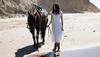 Woman in a white dress and boots walking a horse along the beach