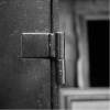 Close-up of a metal door hinge on a partially open textured door, with a blurred window frame in the background. Black and white image.