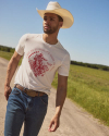 A man wearing a white t-shirt with red print, jeans, and a cowboy hat walks on a rural dirt road beside a green field under a clear blue sky.