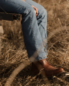 Person in jeans and brown boots sits on a bench in a dry grassy area, holding a yellow object.