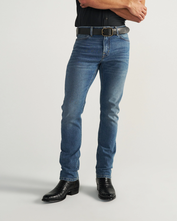 Person wearing blue jeans, a black belt, black shirt, and black cowboy boots stands with arms crossed against a plain background.