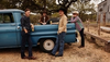 Four men stand and sit around a blue vintage pickup truck in a rural outdoor setting. They are casually dressed in cowboy hats, jeans, and boots and are holding beer cans.