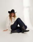 Person with long curly hair wearing a black cowboy hat, white shirt, dark jeans, and black cowboy boots sits on the floor against a plain backdrop.