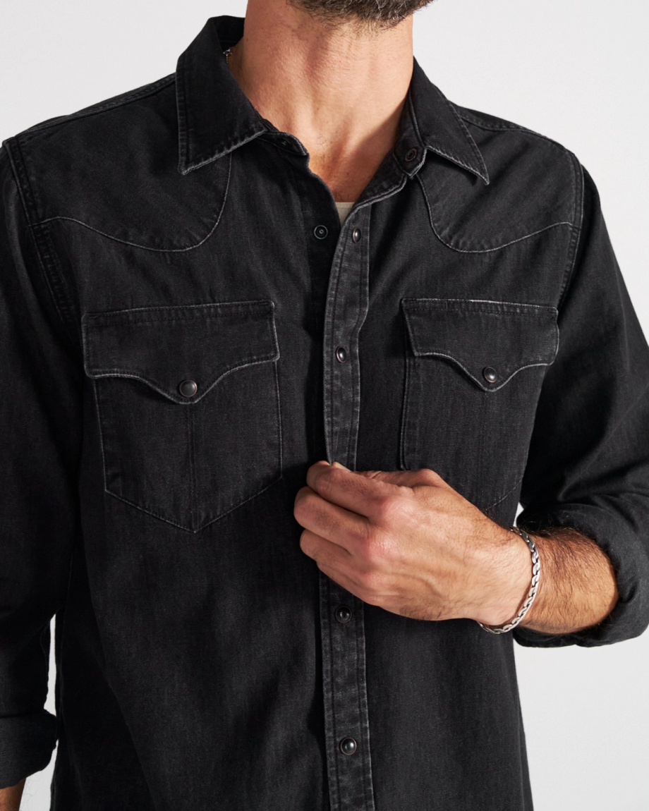 A man wearing a black denim button-up shirt with two chest pockets adjusts the front. Only his torso and part of his face are visible.