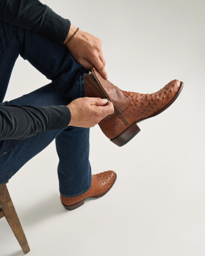 A person wearing blue jeans and a black shirt sits on a chair and zips up The Dean Ostrich, a brown pecan ostrich leather boot.