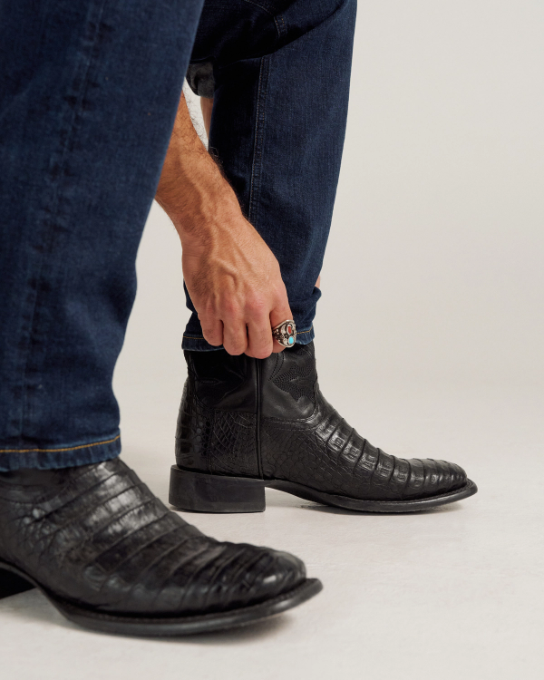 The William in Midnight Caiman. A person wearing blue jeans and The William Caiman black textured leather boots adjusts the zipper on one boot while crouching.