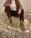 A man wearing a cap, brown jacket, and work boots sits on a pile of rocks in front of a beige backdrop.