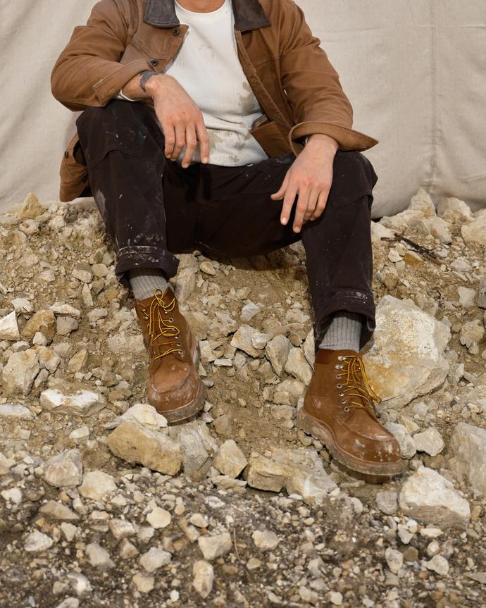A man wearing a cap, brown jacket, and work boots sits on a pile of rocks in front of a beige backdrop.