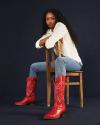 A woman with long curly hair sits sideways on a wooden chair, wearing a white shirt, blue jeans, and red cowboy boots, against a dark background.