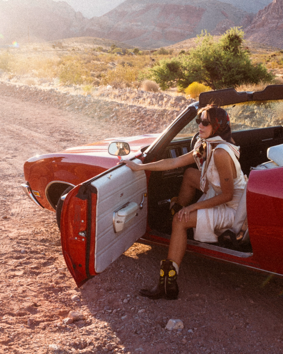 The Jolene in Truffle Cowhide. A person wearing a headscarf and sunglasses sits in a red convertible parked on a dirt road in a desert landscape, with the driver’s door open. Draped across the passenger seat is The Jolene Cowhide, its distinctive pattern catching the sunlight amidst the sandy backdrop.