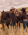 Two people in cowboy hats stand beside saddled horses in a grassy field under a cloudy sky.