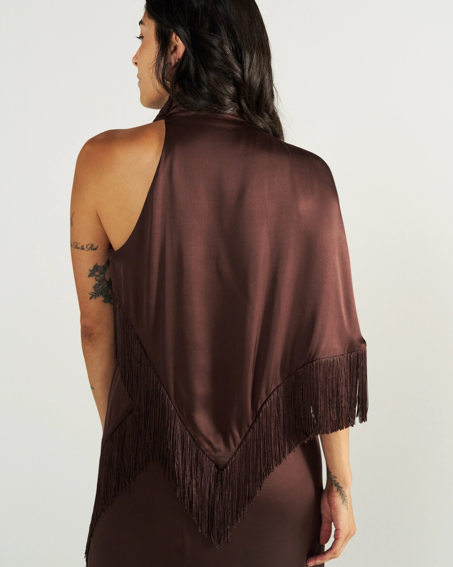 Woman in a sleeveless brown dress with a fringed shawl draped over her back, facing away from the camera against a plain background.