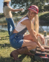 A woman in overalls and a red cap crouching next to a tackle box near a river, while a person in the background, wearing a cowboy hat, casts a fishing line.