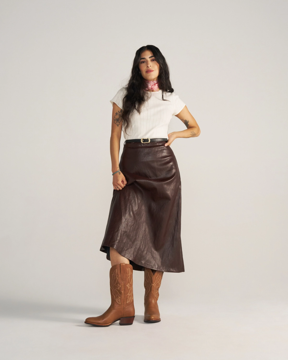 A woman stands against a plain background wearing a white short-sleeve top, brown leather skirt, brown belt, and brown cowboy boots—a chic look inspired by The Britt Goat’s signature latte tones.