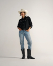 The Britt in Midnight Cowhide. A woman stands against a plain background wearing a white cowboy hat, black shirt, blue jeans, and striking Midnight Cowhide boots.