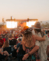 A couple wearing hats enjoys a sunset concert outdoors, embracing while facing a lively crowd and stage.