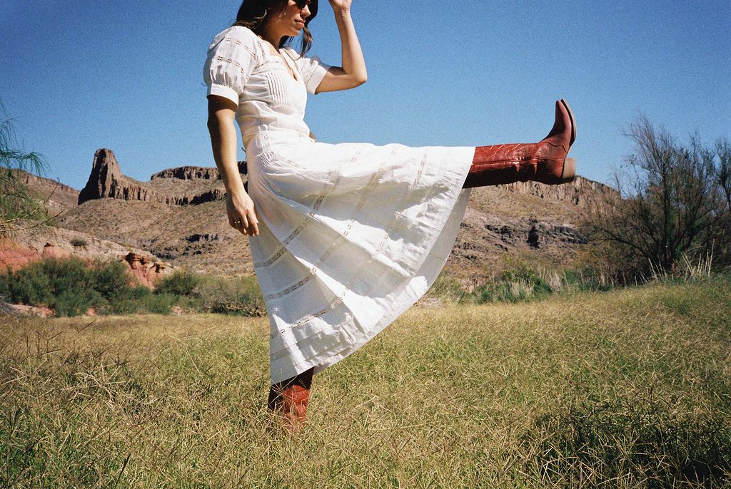Person in a long white dress and red boots kicking the air in a grassy field with mountains in the background.