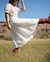 Person in a long white dress and red boots kicking the air in a grassy field with mountains in the background.