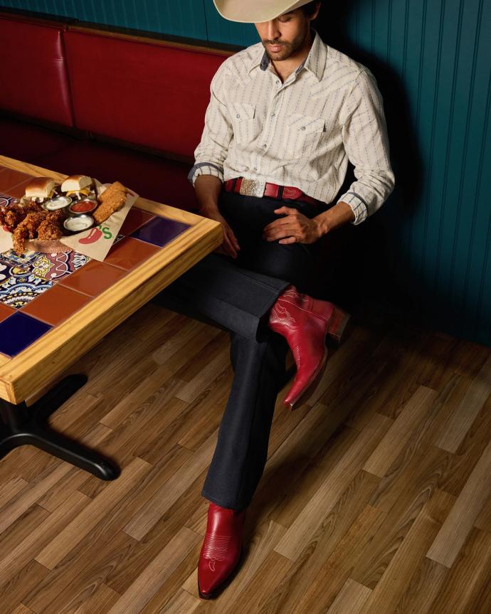 A man in a cowboy hat, patterned shirt, and red boots sits with legs crossed at a tiled table with fried food and dipping sauces.