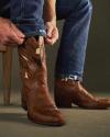 A person wearing blue jeans sits on a wooden chair and pulls on a brown leather cowboy boot with decorative stitching.
