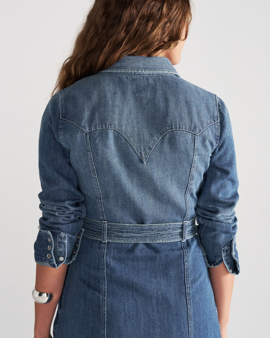 A person seen from behind wearing a blue denim shirt dress with a Western-style yoke and a belted waist.