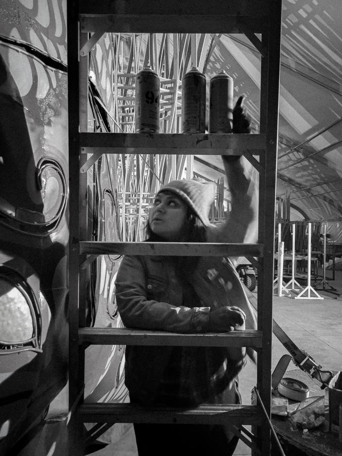 A woman in a beanie arranges cans on a shelf in a warehouse, with a focus on her face framed by the shelf's structure and surrounding objects.