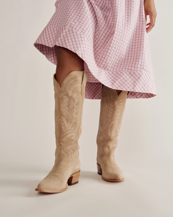 A person wearing a pink checkered dress and beige knee-high cowboy boots stands against a plain background.