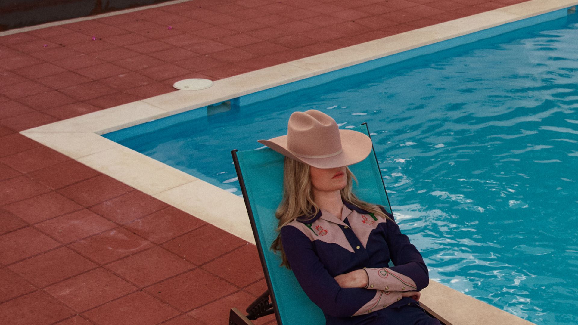 A person in a cowboy hat and western shirt lounges with arms crossed on a turquoise pool chair beside a swimming pool with red tile flooring.