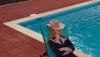 A person in a cowboy hat and western shirt lounges with arms crossed on a turquoise pool chair beside a swimming pool with red tile flooring.