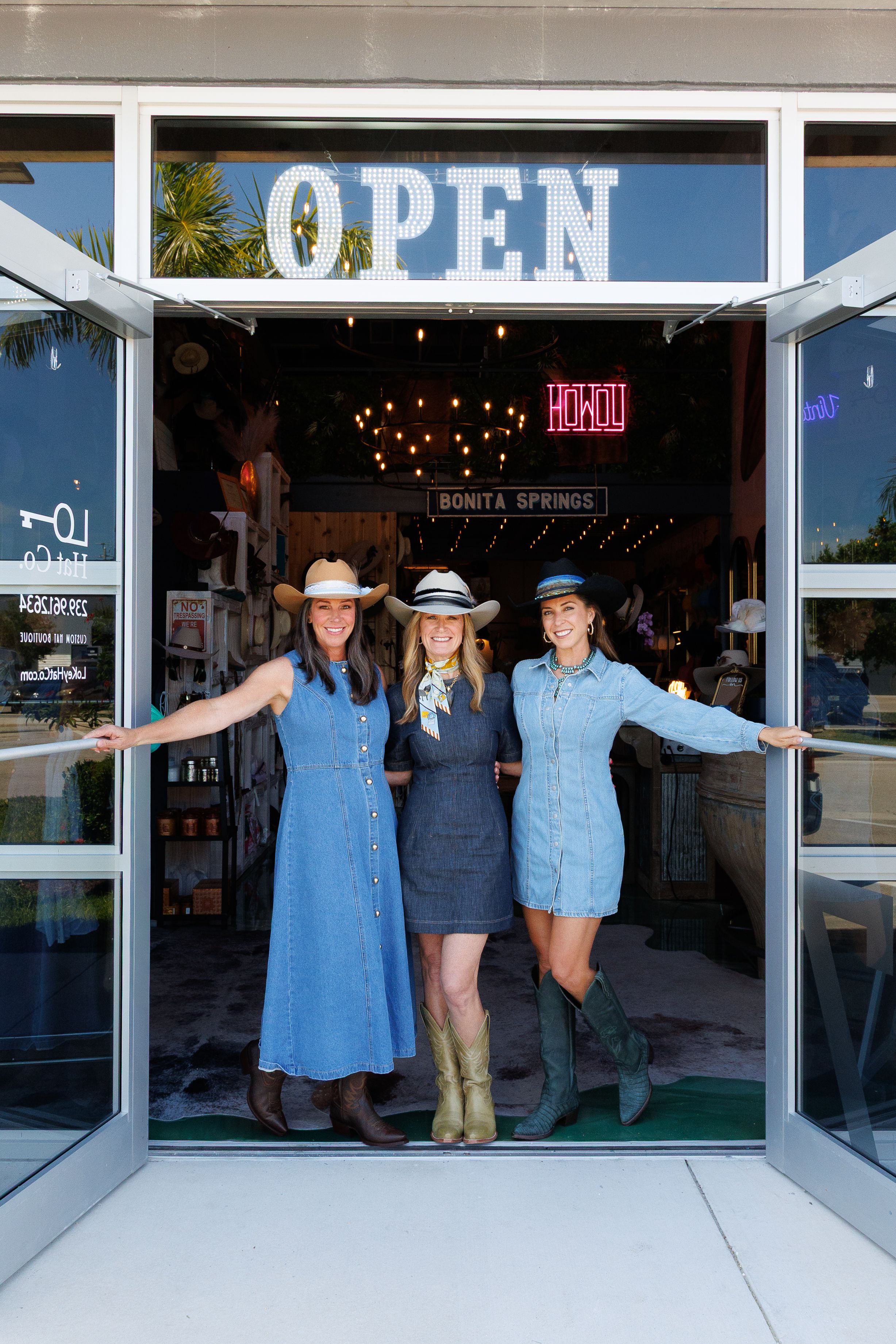 Three women wearing denim and cowboy hats stand in the open doorway of a store with a lit "OPEN" sign above them.