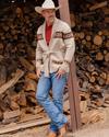 A man in a cowboy hat and patterned cardigan sweater stands leaning against a wooden post in front of a large stack of firewood. He is wearing jeans and boots.