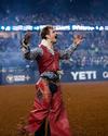 A rodeo rider in decorative chaps and a patterned shirt stands on a dirt arena, arms raised, with a cheering crowd in the background.