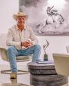 A person in a cowboy hat sits in a chair with a horse photo in the background and a trophy on the table.