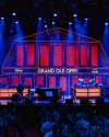 A stage illuminated with blue and red lights features a barn-like backdrop displaying "Grand Ole Opry." People are seated in front, watching performers on stage with musical instruments.