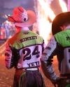 A rodeo rider wearing a white hat and a green, black, and white jersey with "KIMZEY 24 YETI" stands near another rider as flames shoot up in the background.