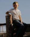 A boy in a white T-shirt and jeans sits on a wooden fence outdoors under a clear blue sky.