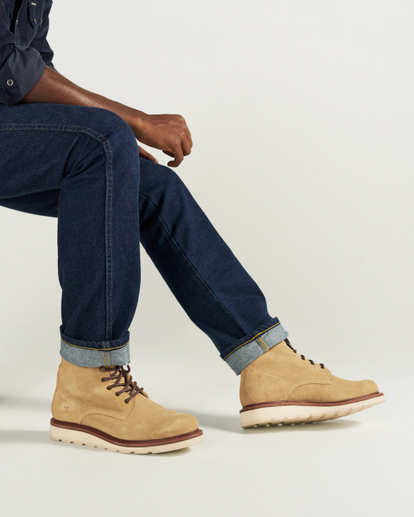 The Knox Lace Up Shearling in Sandstone Roughout. Person sitting on a stool wears dark blue jeans with cuffed hems and tan Sandstone Roughout lace-up boots against a plain, light background.
