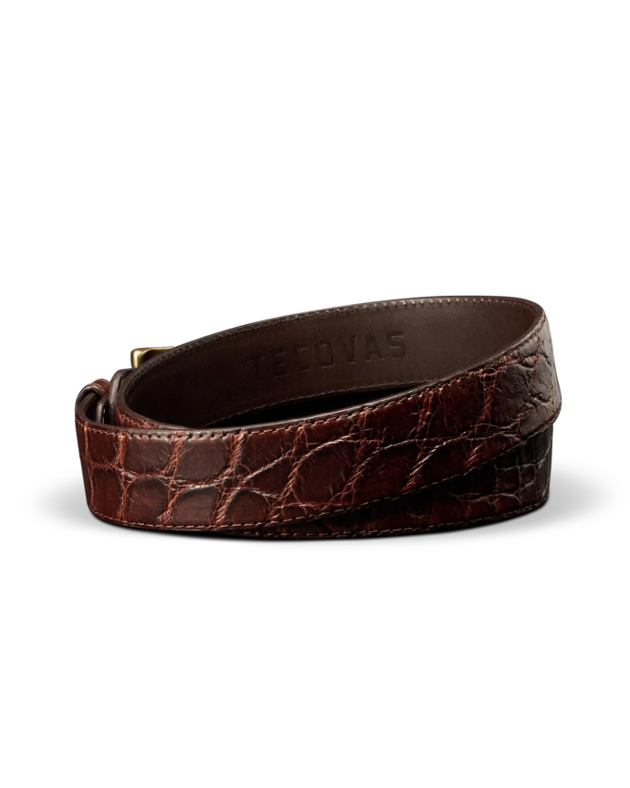 A brown crocodile-embossed leather belt with a gold buckle, coiled on a white background. The brand name “Tecovas” is stamped inside.
