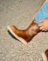 A person wearing blue jeans puts on a brown leather boot while standing on gravel.