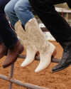 Three people sitting on a metal fence, wearing brown, white, and black cowboy boots, with a dirt ground in the background.