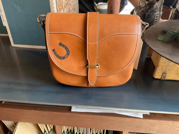 A brown leather crossbody bag with a horseshoe emblem on the front is displayed on a table.