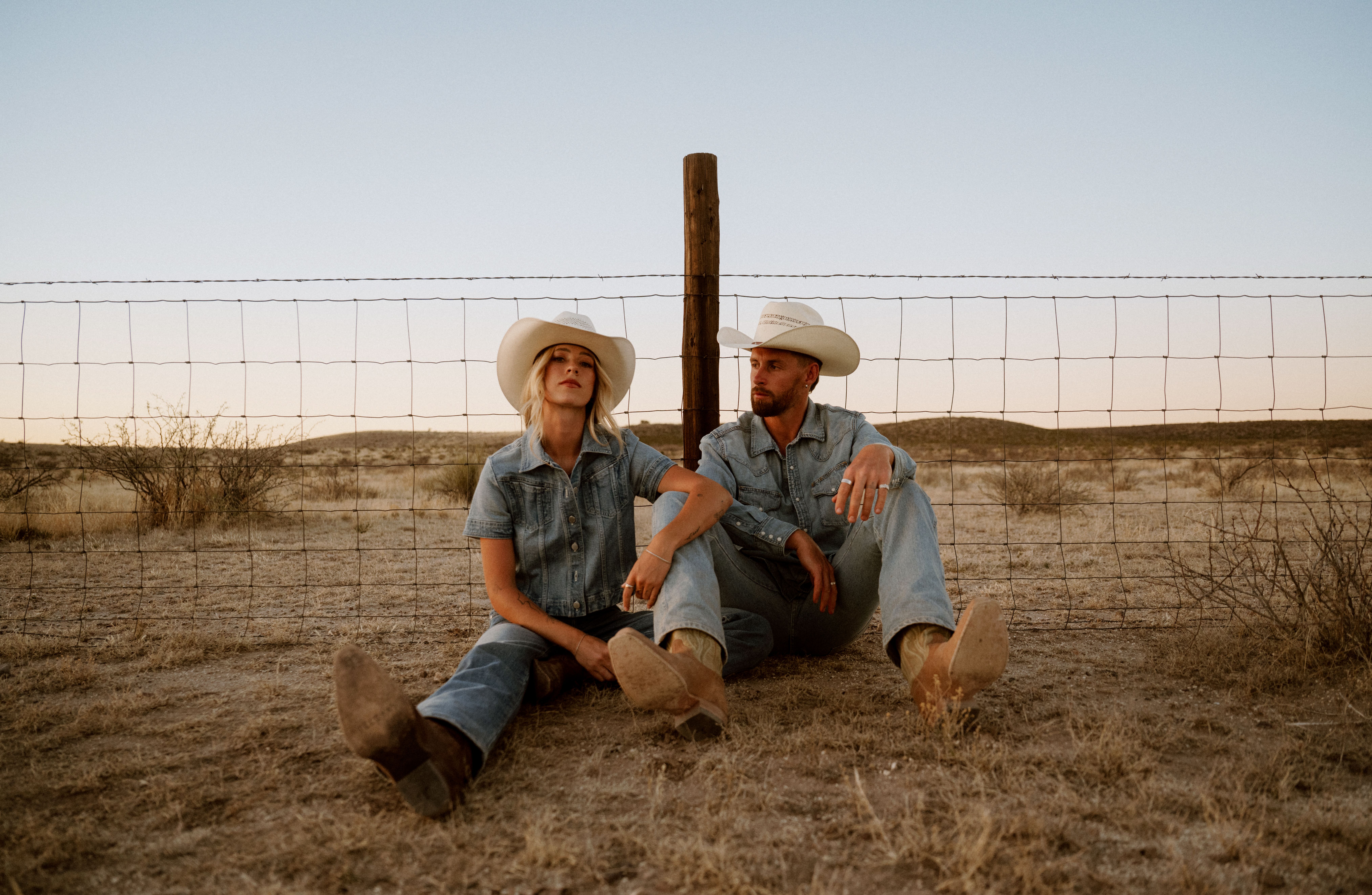 Two people in cowboy hats and denim clothing sit on the ground by a wire fence in a dry, open landscape at sunset.
